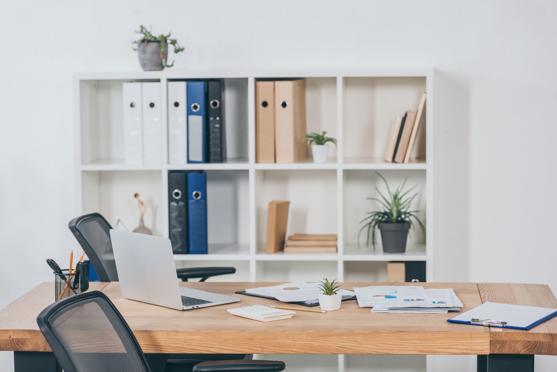 table in office with laptop, documents, chairs and bookcase fo folders on background